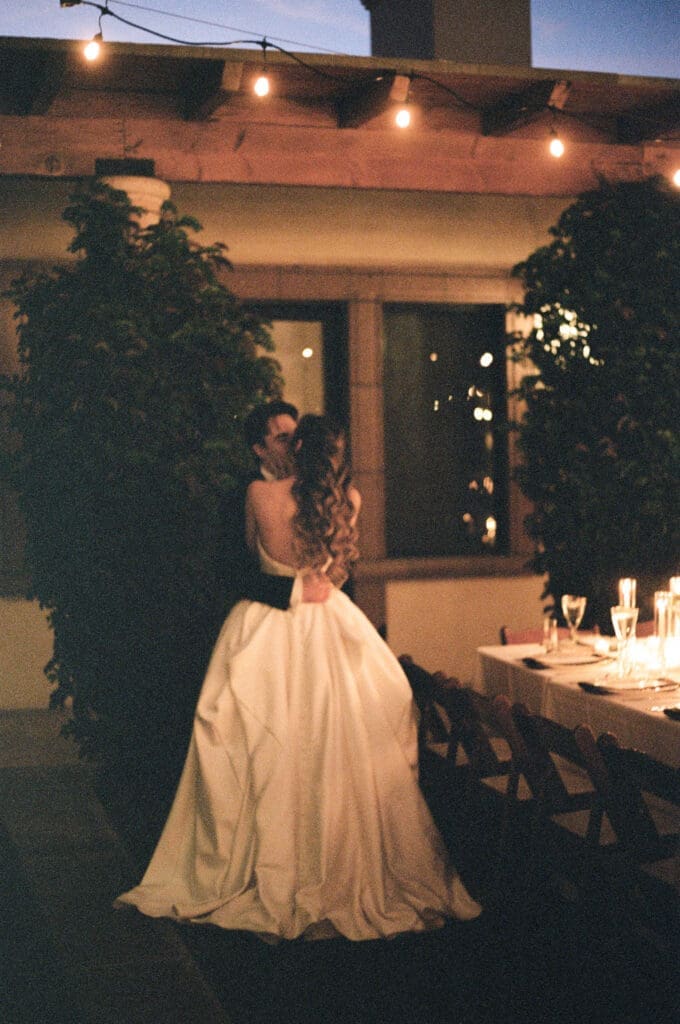 Bride and groom embracing beside a candlelit dinner table under string lights at dusk.