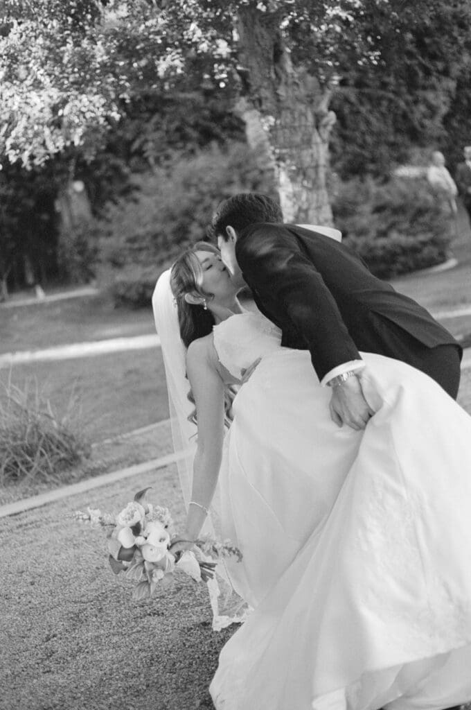 Black and white photo of the bride and groom kissing outdoors, her bouquet resting on the ground.