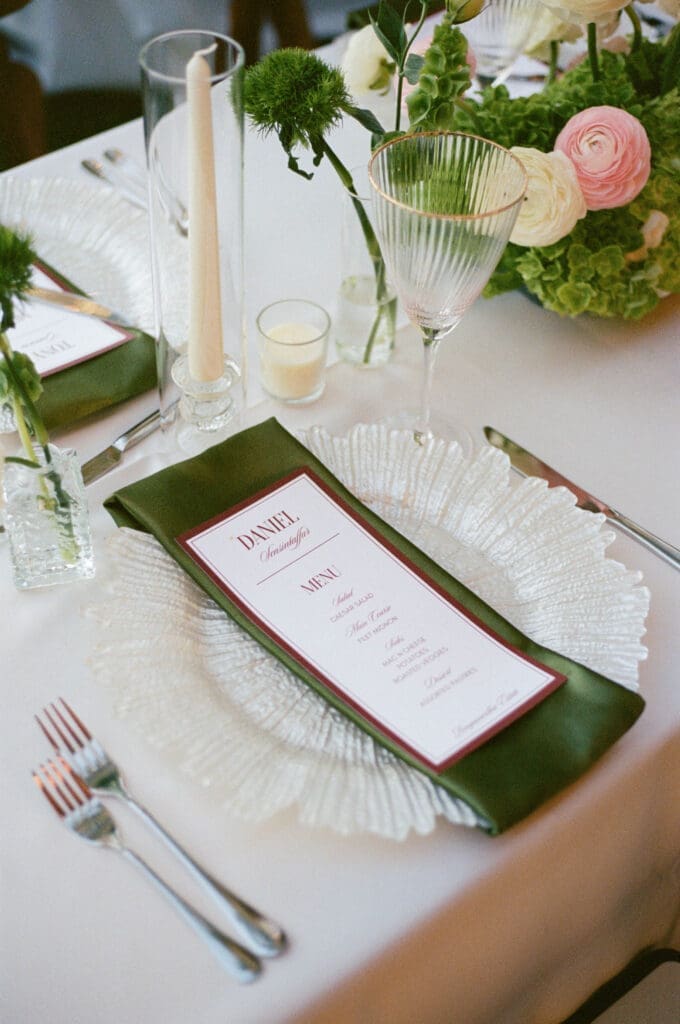Reception table setting detail with a green menu card, taper candle, and floral accents.