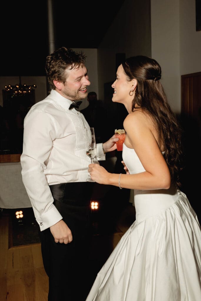 Bride and groom laughing together while holding drinks during the reception, cozy indoor lighting