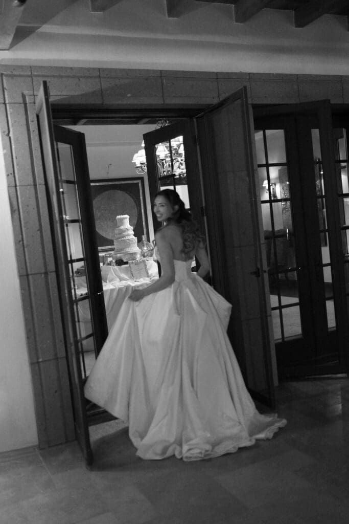 Black and white photo of the bride walking through a doorway toward the cake table.