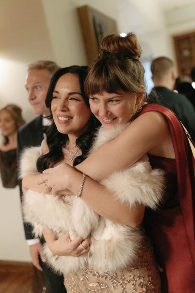 Two guests in fur stoles embracing and laughing at a lively moment during the reception.