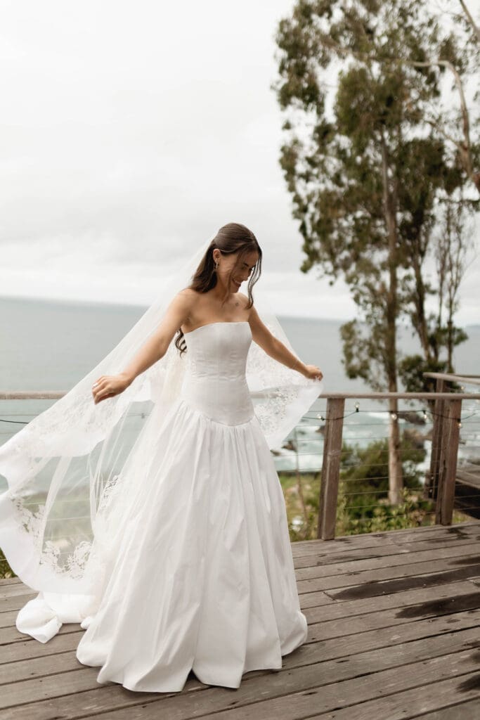 Bride twirling on a wooden deck overlooking the ocean, her veil caught in the coastal wind.
