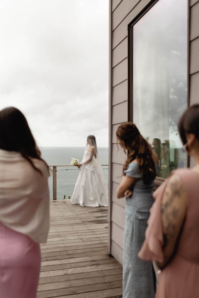 Bridesmaids first look on a wooden deck toward the bride, with the Pacific Ocean visible beyond the railing.
