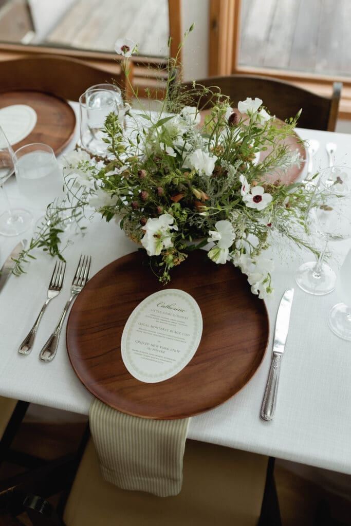 Place setting detail with a wood charger plate, menu card, and white floral arrangement.