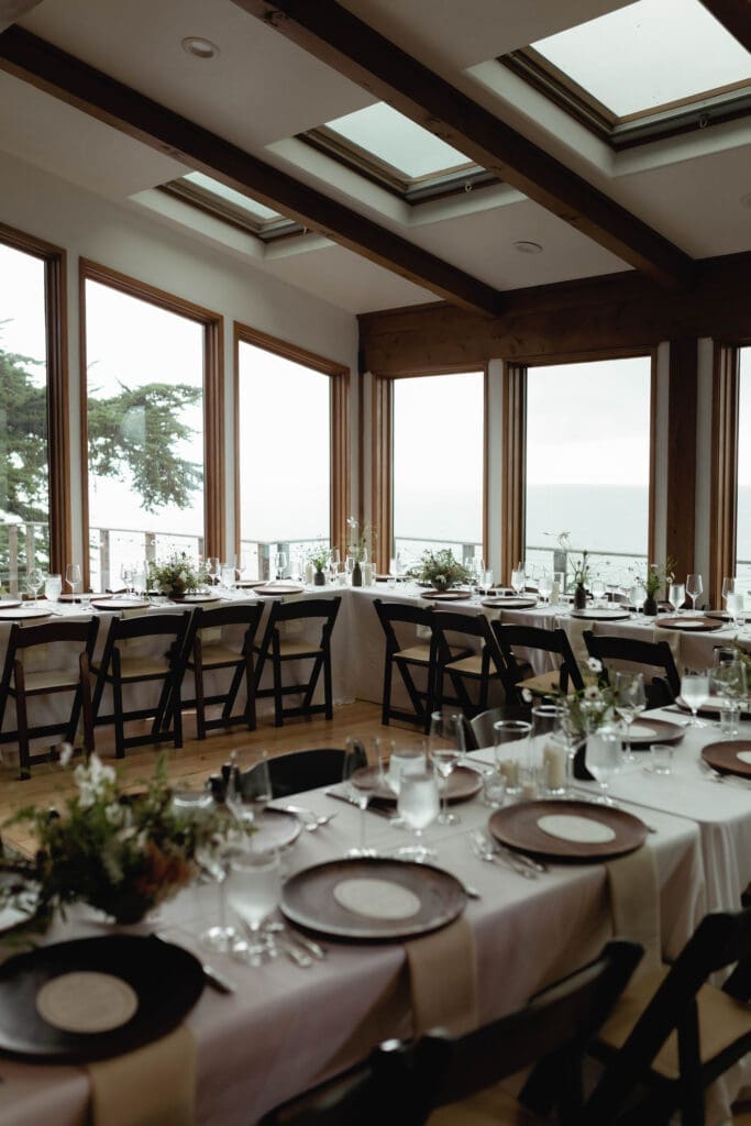 Long candlelit dinner table set beneath skylights and floor-to-ceiling windows during the intimate indoor reception at Wind and Sea Estate.