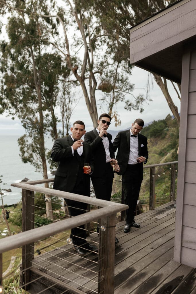 Groomsmen standing on a wooden deck under an overcast Big Sur sky, ocean visible below.