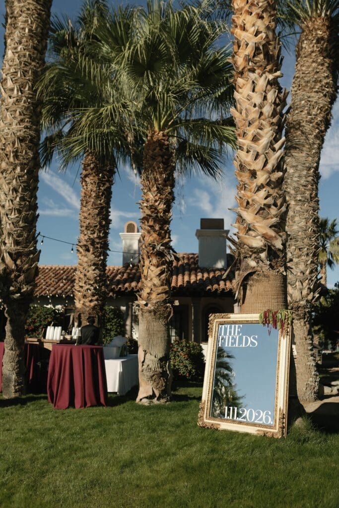 Framed welcome sign and bar setup nestled under palm trees at a Coachella Valley wedding reception.