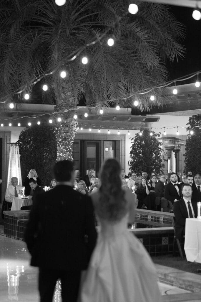 Black and white photo of the bride and groom walking into their reception under string lights as guests cheer.