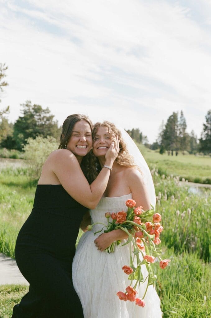 Bride laughing while embracing a bridesmaid in a black dress, holding a coral tulip bouquet in a grassy meadow