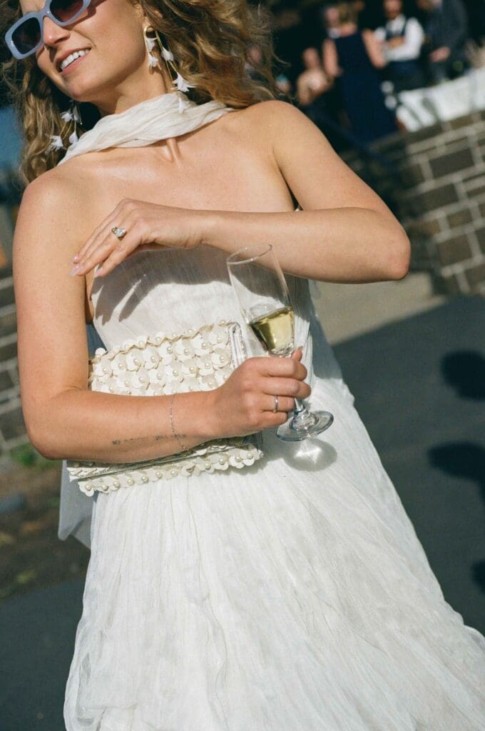Bride wearing sunglasses and a floral headscarf, smiling and holding a champagne flute at her reception.