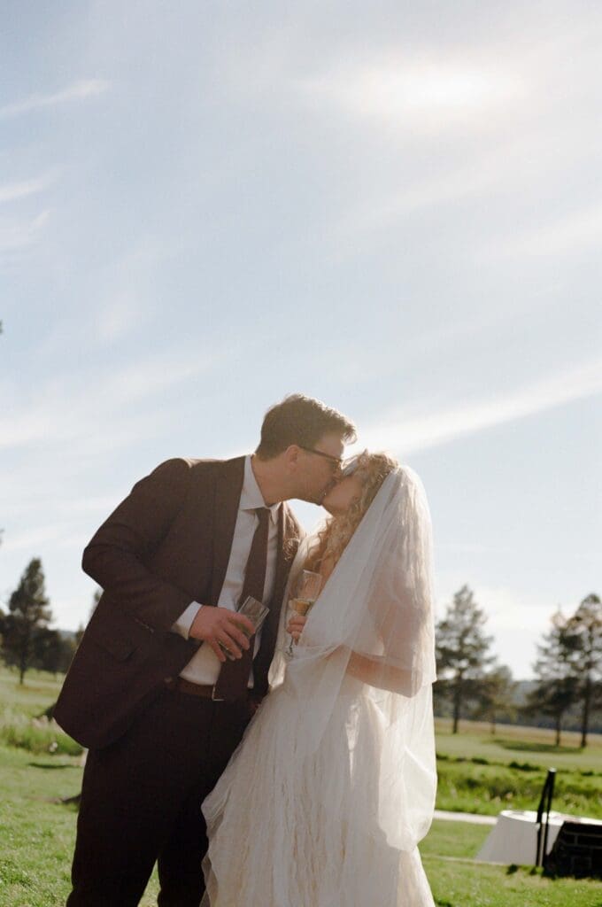Bride and groom sharing a kiss outdoors at their wedding, holding champagne glasses under a wide open sky.