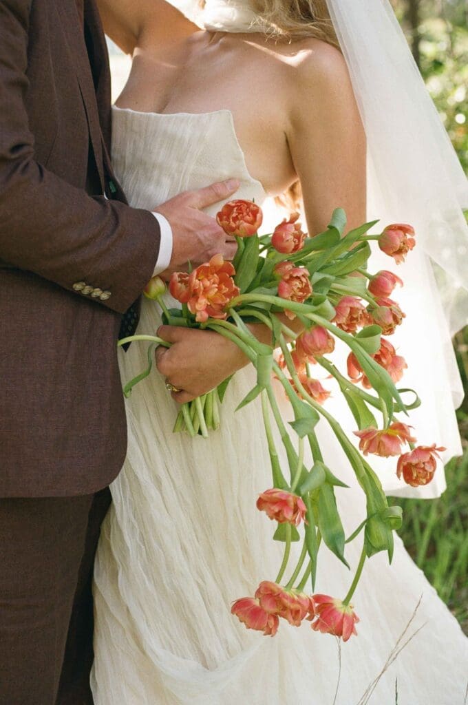 Close-up of a bride's textured wedding dress and coral tulip bouquet, with the groom's hand at her waist.