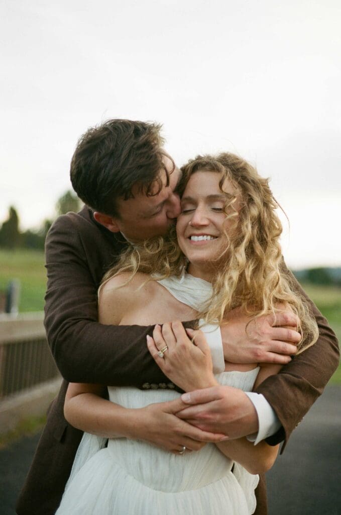 Groom kissing the bride's cheek while embracing her outdoors in soft afternoon light.