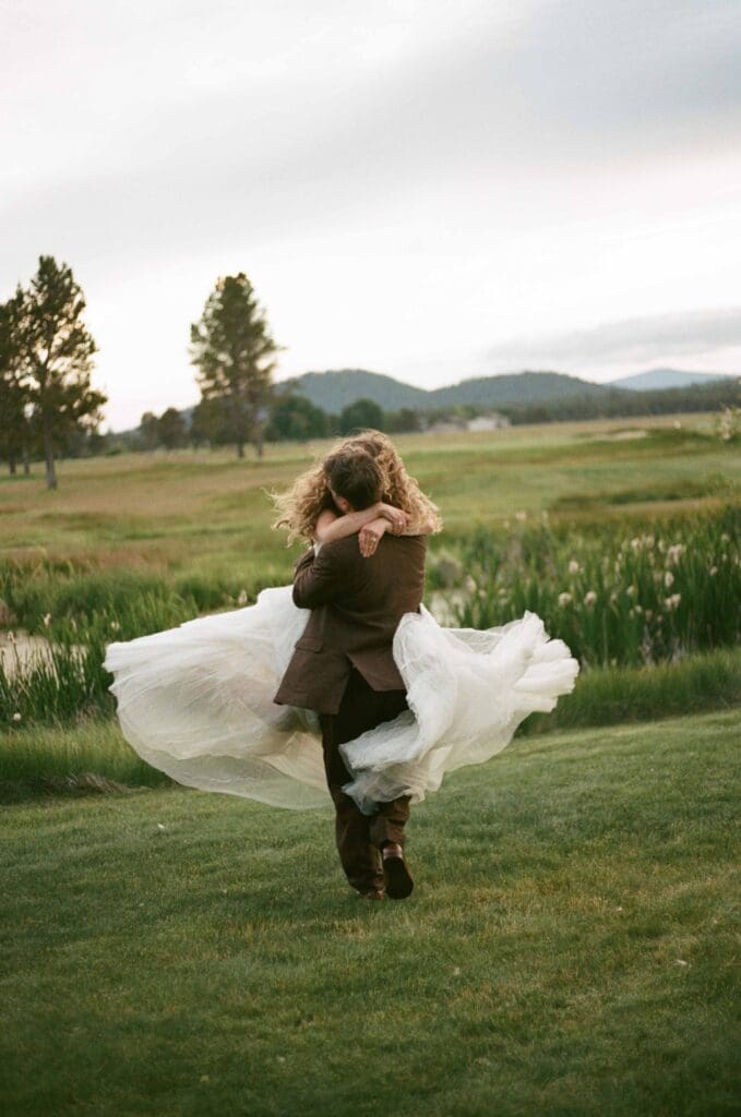 Film wedding photography of a groom carrying his bride across an open meadow, her dress and veil caught mid-motion.