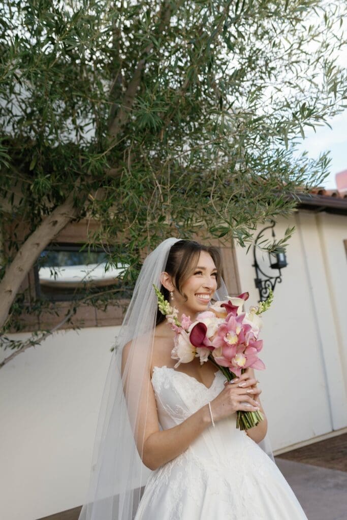 Bride smiling and holding her bouquet beneath an olive tree at her Bougainvillea Estate wedding.