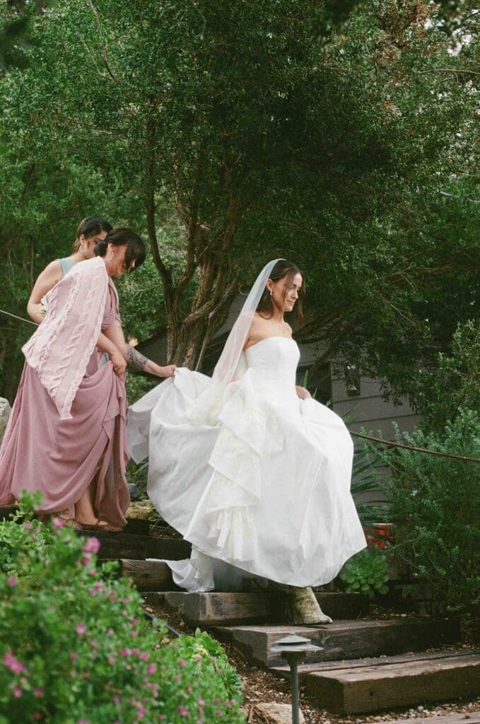 Bridesmaids helping the bride down stone garden steps, surrounded by greenery