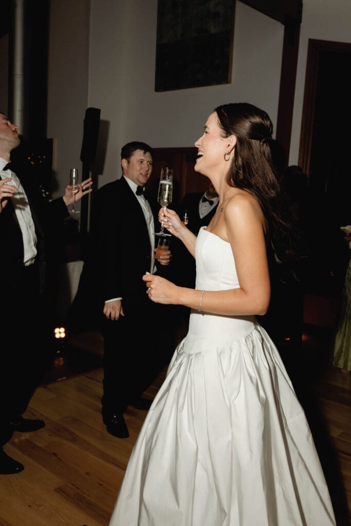 Bride laughing and dancing with a glass of champagne, guests celebrating around her