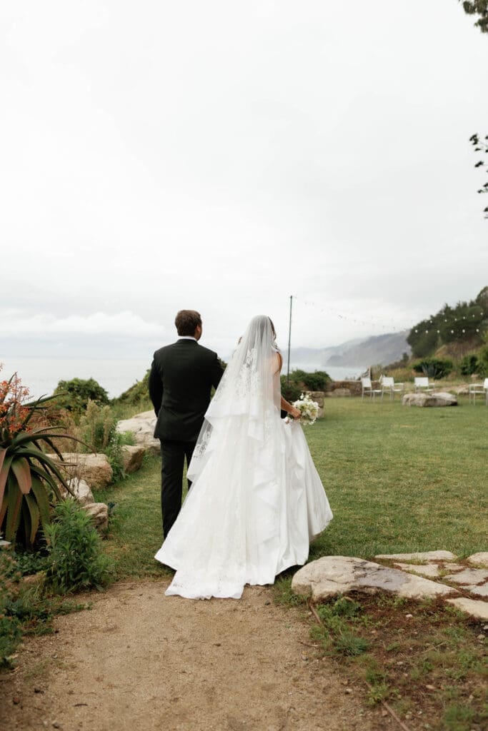 Bride and groom walking hand in hand toward the lawn at Wind and Sea Estate, the ocean visible in the distance.