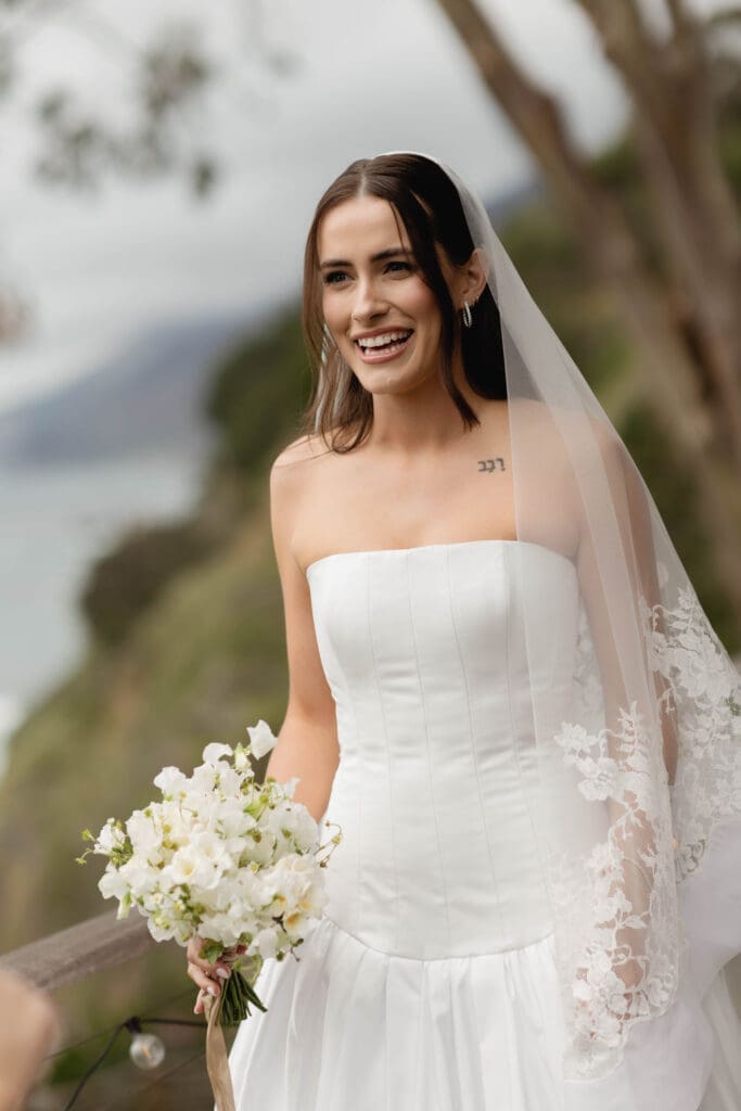 Bride smiling and holding a white bouquet, misty coastal hills behind her on an overcast Big Sur morning.