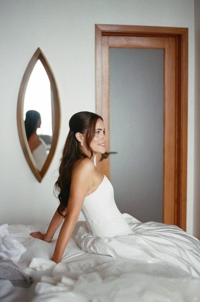 Bride sitting on the bed in her wedding gown during quiet getting-ready moments, reflected in an oval mirror.