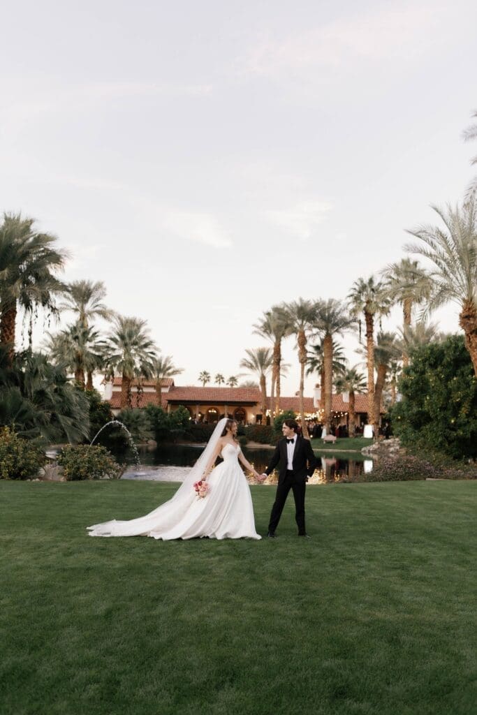 Bride and groom holding hands on a palm-lined lawn beside a pond at their Bougainvillea Estate wedding.