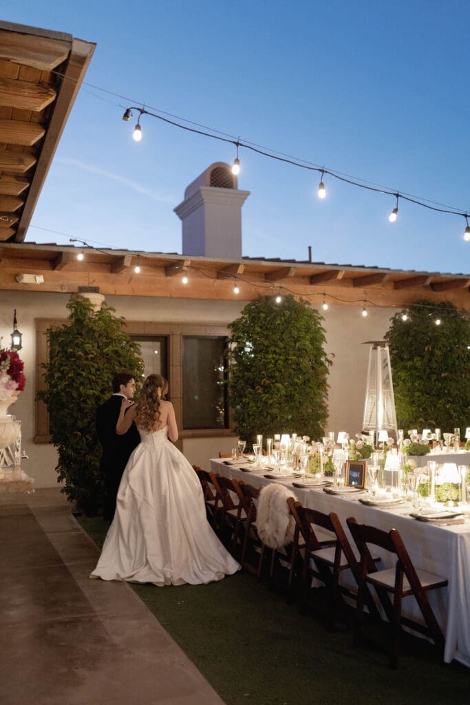 Bride and groom standing beside a candlelit outdoor dining table at Bougainvillea Estate, string lights and the estate's chimney overhead.