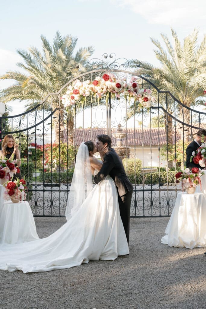 Bride and groom sharing their first kiss in front of a floral-wrapped iron gate at their outdoor ceremony."