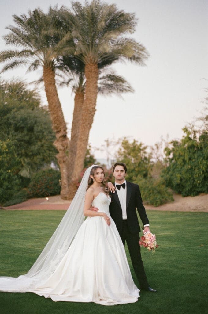 Bride and groom posing together on the lawn at Bougainvillea Estate, tall palms rising behind them at dusk.