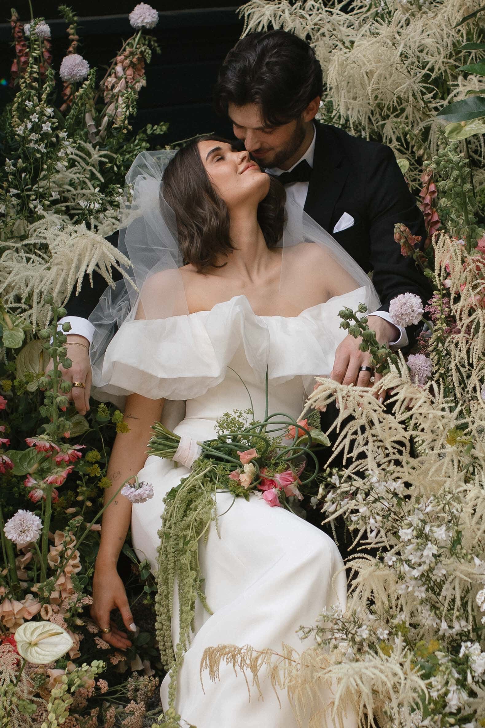 Bride and groom embracing among lush dried florals, bride in an off-shoulder gown and veil, groom in a black tuxedo — the hero image for a film vs digital wedding photography feature.