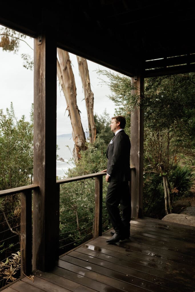 Groom standing alone on a covered wooden porch overlooking the ocean