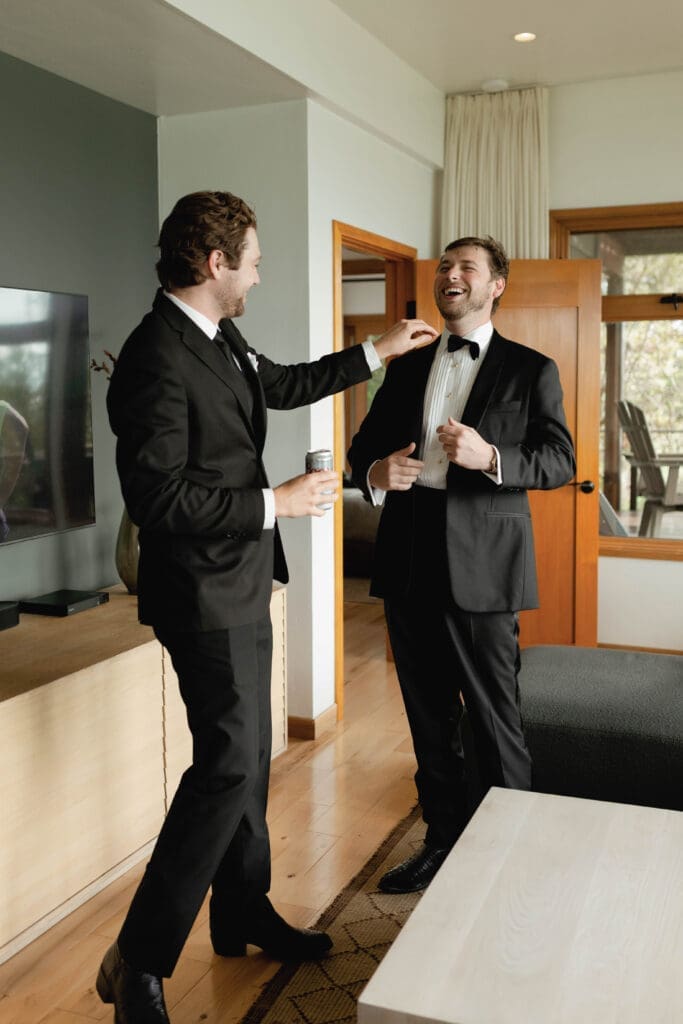 Groomsman laughing while helping the groom adjust his collar during getting-ready moments indoors.