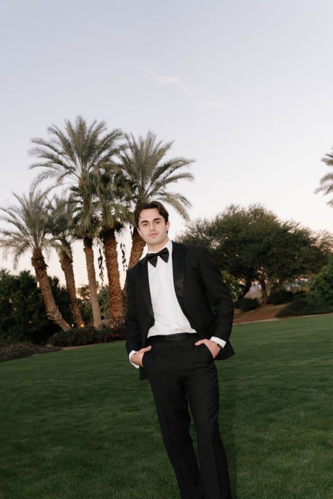 Groom in a black tuxedo posing on the lawn with palm trees in the background.