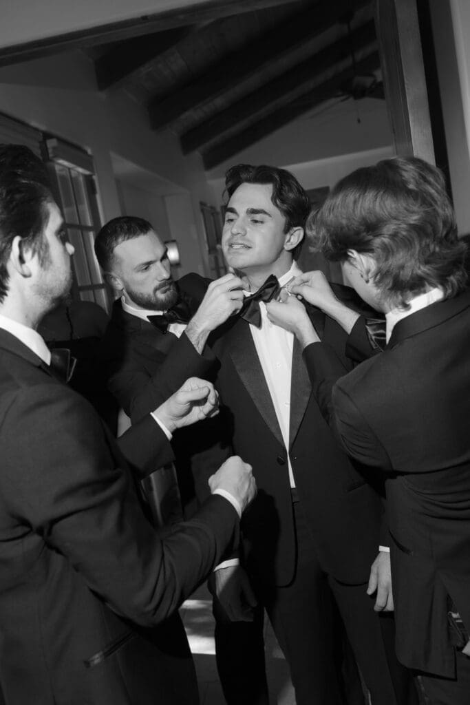 Black and white candid of groomsmen helping the groom fix his bowtie before the ceremony.