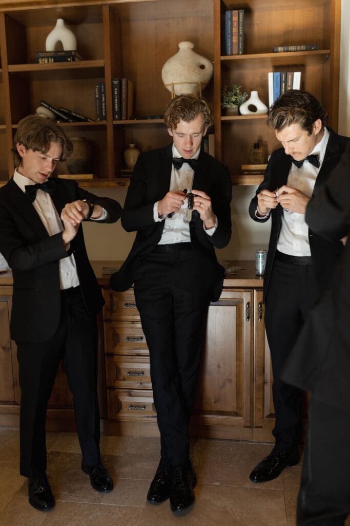 Groomsmen in tuxedos adjusting cufflinks and watches in a wood-paneled room.