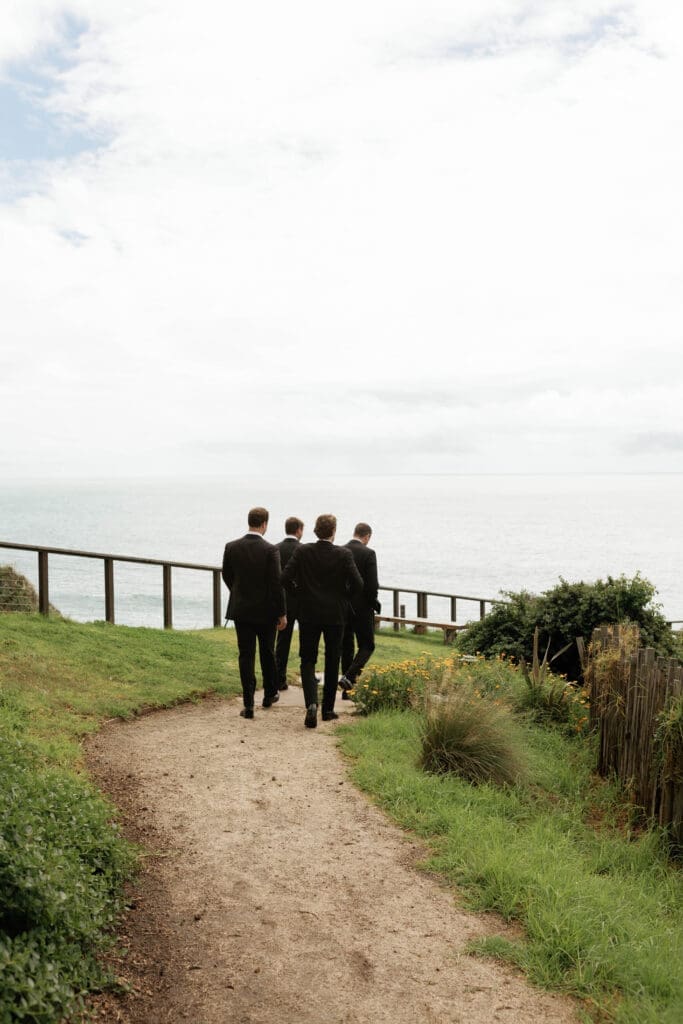 Groomsmen walking together along a cliffside path overlooking the ocean.