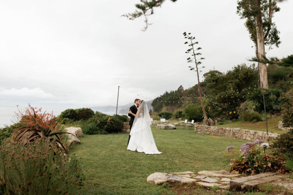 Bride and groom embracing on the cliffside lawn at Wind and Sea Estate during a rare clear-sky window in their otherwise rainy Big Sur wedding.