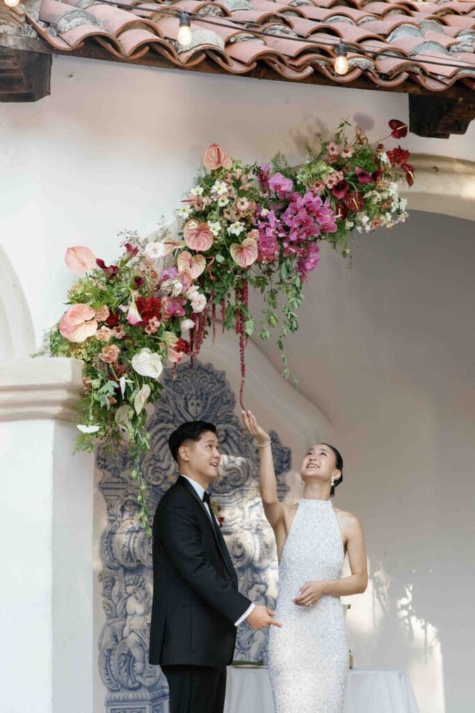 Digital wedding photography of a couple at their ceremony beneath a dramatic hanging floral installation under a tiled archway