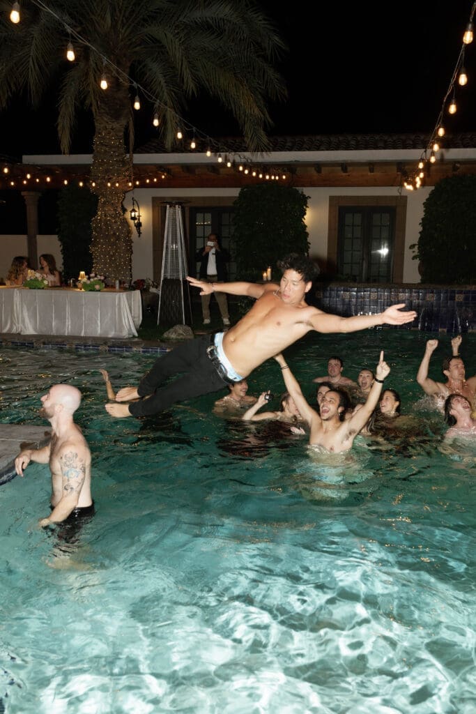 Guest being tossed into the pool as others cheer, string lights and palm trees overhead in a high energy end-of-night moment.