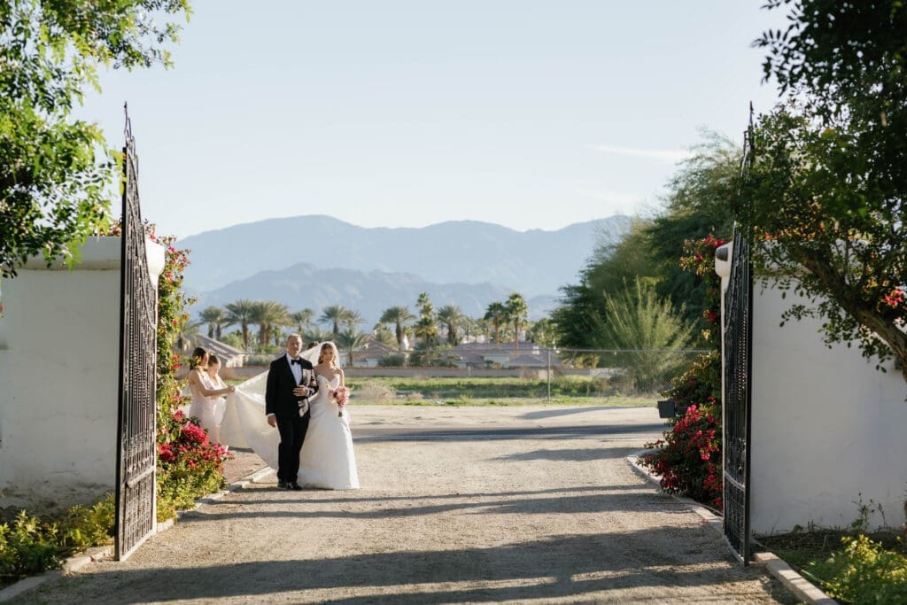 Bride and her father walking through Bougainvillea Estate's iron entrance gate, with Coachella Valley mountains and real bougainvillea blooms framing the drive.