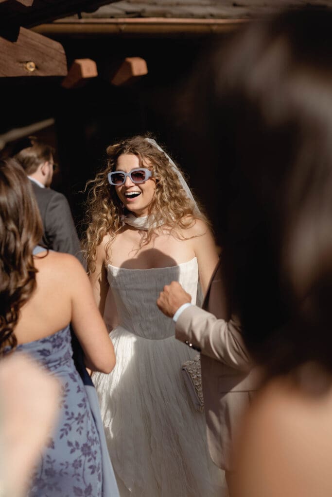 Bride wearing sunglasses and laughing with guests at the entrance of a rustic timber building