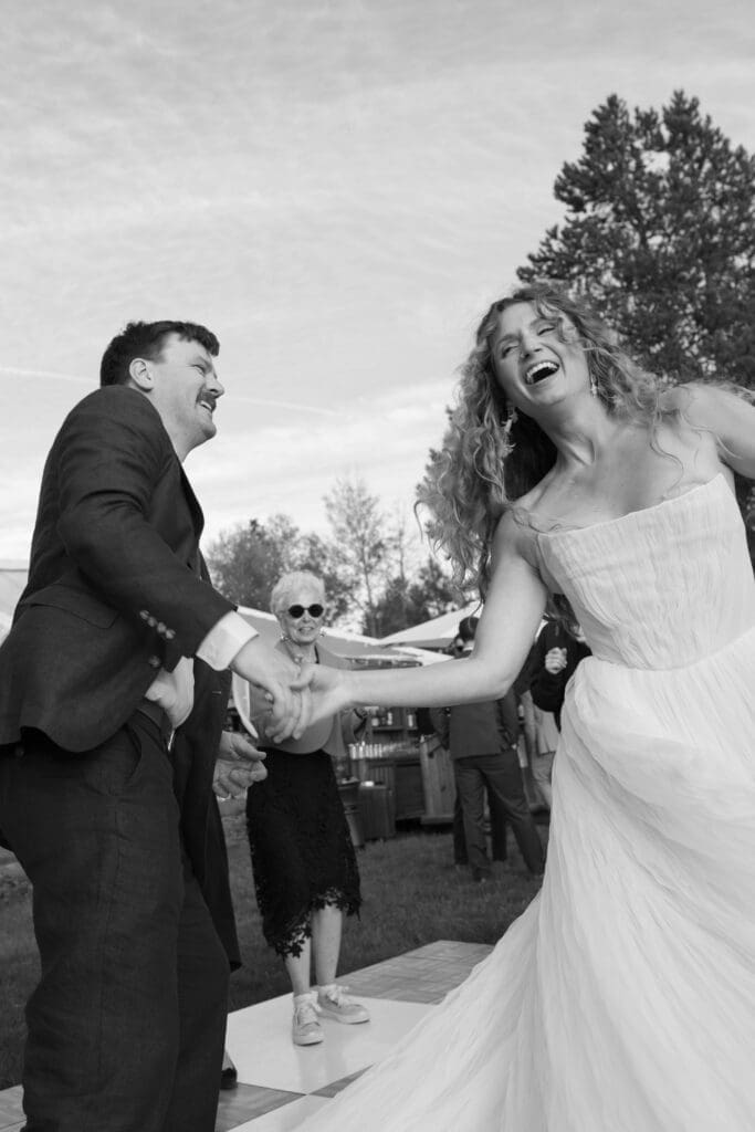 Black and white photo of the bride and groom laughing and holding hands while dancing outdoors.