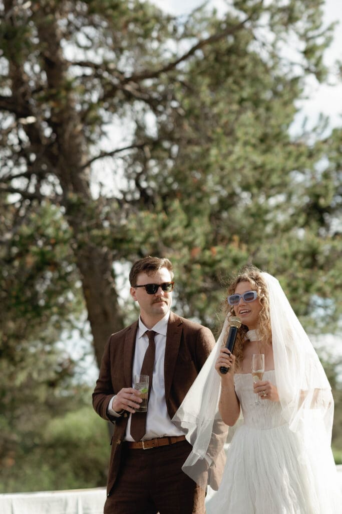 Bride in sunglasses holding a microphone next to the groom during an outdoor toast under tall pines.