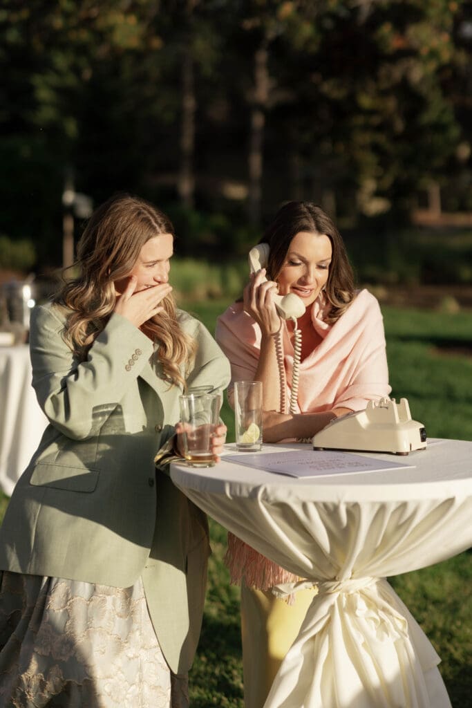 Wedding guests laughing while leaving a message on a vintage rotary phone guest book at an outdoor cocktail table.