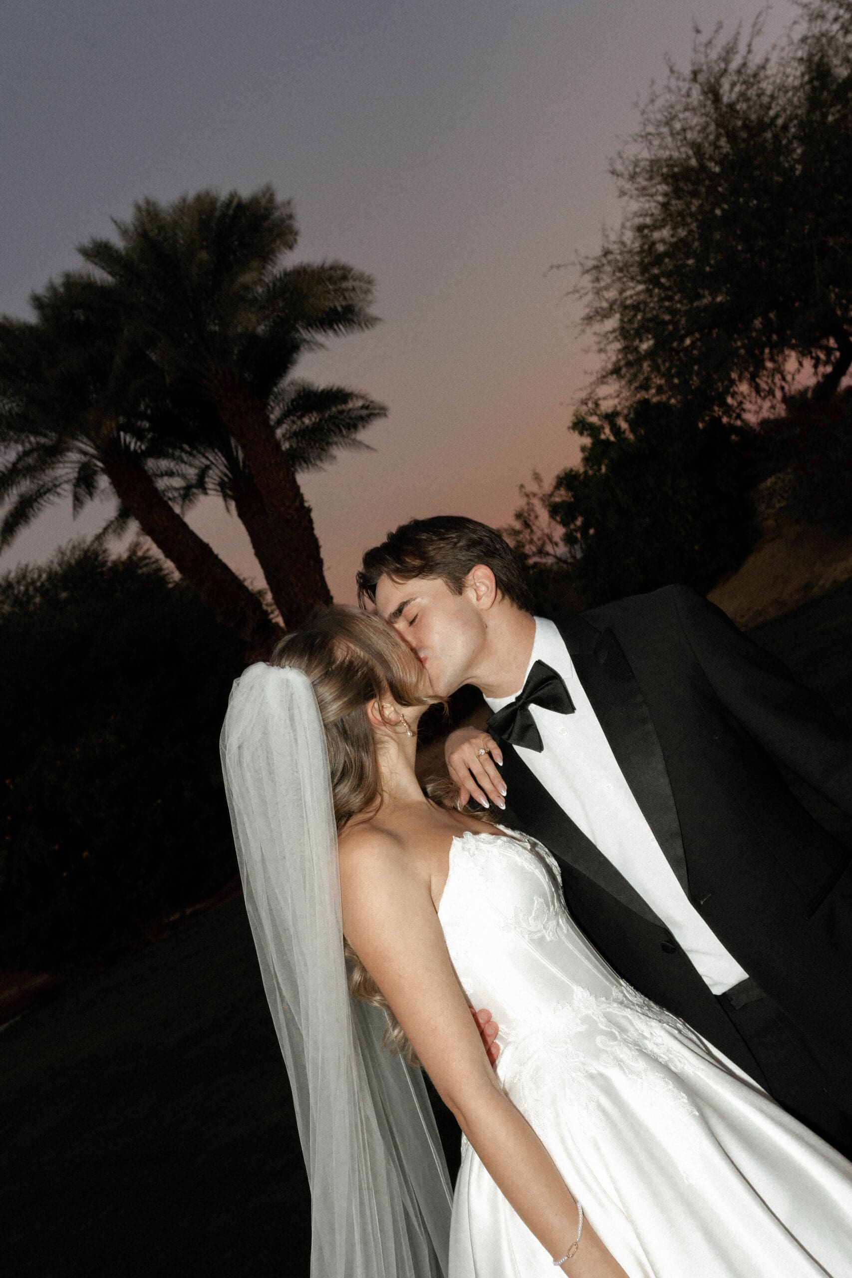 Groom kissing his bride's temple against a dusky sky with palm trees behind them.