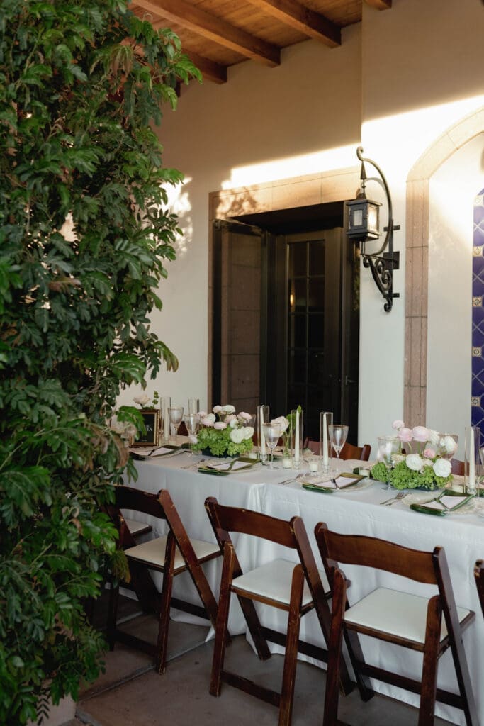 Reception table set beneath a covered patio with string lights and lush greenery.