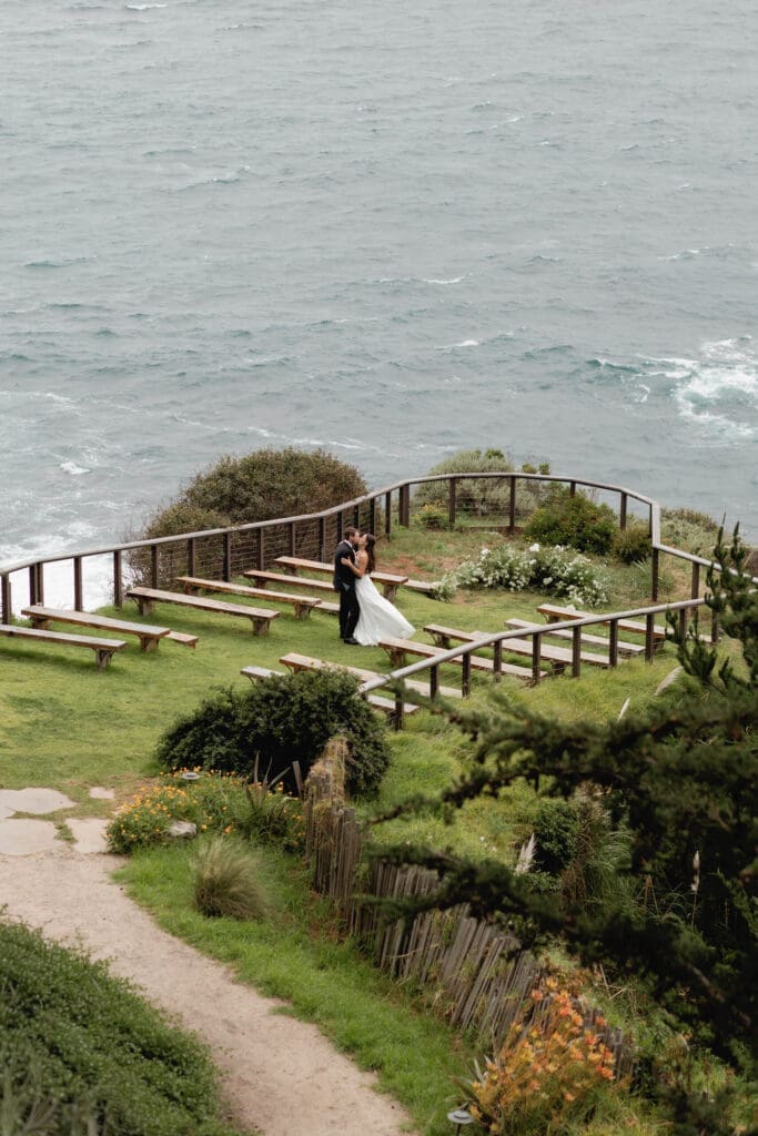 Bride and groom kissing at the cliffside ceremony amphitheater at Wind and Sea Estate, overlooking the churning Pacific during their brief window of clear skies.