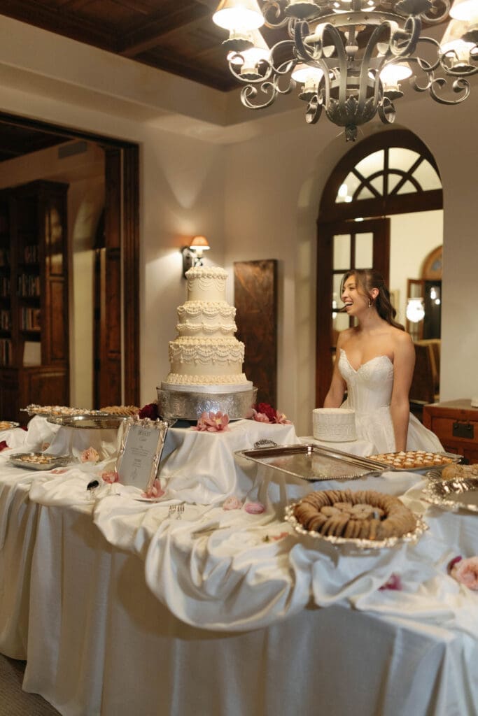 Bride laughing beside the dessert table and tiered wedding cake under a chandelier.