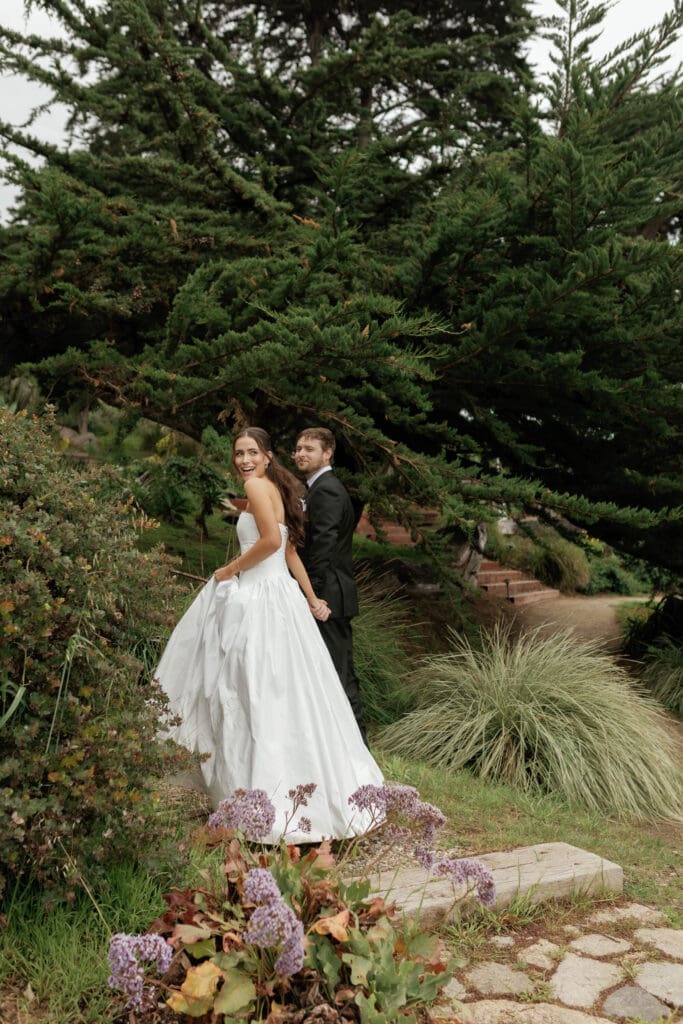 Bride glancing back and laughing while walking hand in hand with the groom through lush garden greenery.