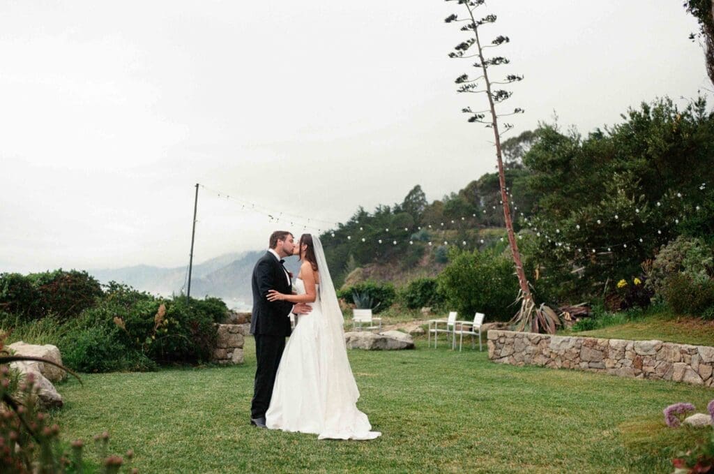 Bride and groom kissing on the cliffside lawn at Wind and Sea Estate, string lights and coastal mountains behind them."
